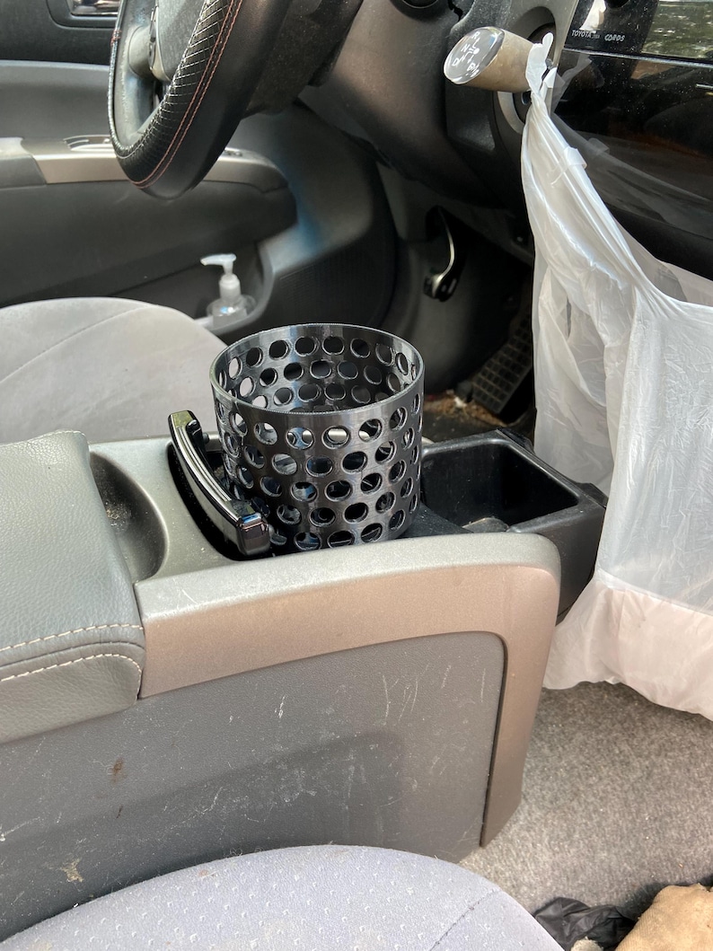 Puede incluir: Un portavasos de metal negro con un dise&ntilde;o perforado se encuentra en la consola central de un coche. El portavasos es cil&iacute;ndrico y tiene numerosos agujeros peque&ntilde;os. El interior del coche incluye un volante, asientos y una bolsa de pl&aacute;stico.