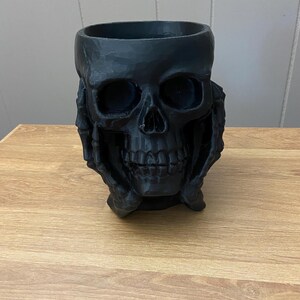 Peut inclure: Un pot en forme de cr&acirc;ne noir avec une large ouverture. Les traits du cr&acirc;ne, y compris les orbites, le nez et les dents, sont clairement d&eacute;finis. Le pot est tenu par deux mains. L'objet est fait d'un mat&eacute;riau sombre et repose sur une surface en bois.