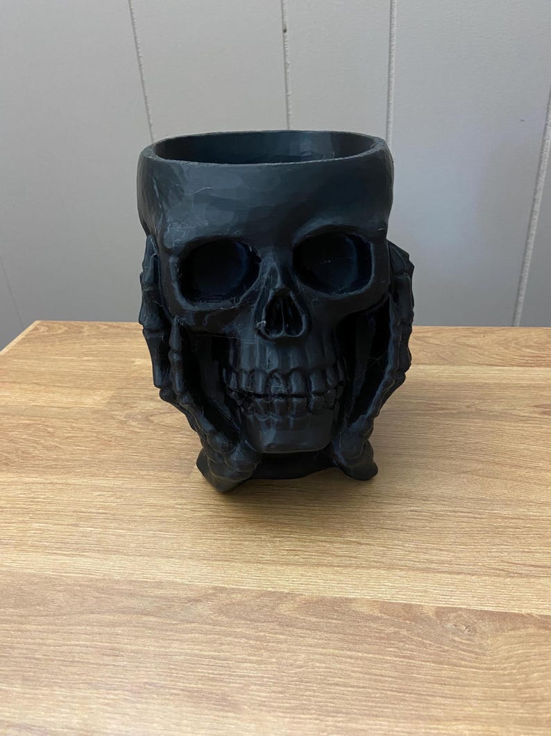 Peut inclure: Un pot en forme de cr&acirc;ne noir avec une large ouverture. Les traits du cr&acirc;ne, y compris les orbites, le nez et les dents, sont clairement d&eacute;finis. Le pot est tenu par deux mains. L'objet est fait d'un mat&eacute;riau sombre et repose sur une surface en bois.