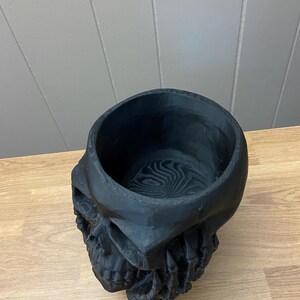 Peut inclure: Un pot en forme de cr&acirc;ne noir avec un dessus &eacute;vid&eacute;. Le pot est fait d'un mat&eacute;riau sombre et pr&eacute;sente des traits faciaux d&eacute;taill&eacute;s, dont une m&acirc;choire et des orbites visibles. L'int&eacute;rieur pr&eacute;sente un motif tourbillonnant.