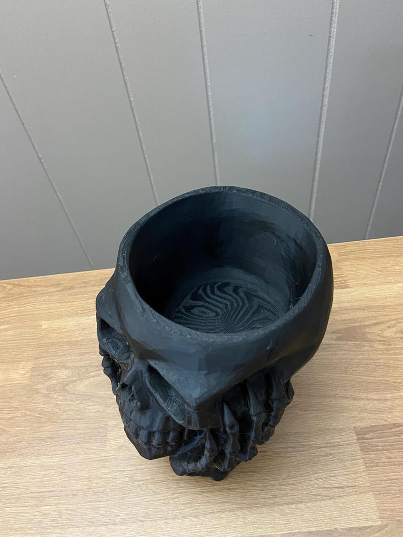 Peut inclure: Un pot en forme de cr&acirc;ne noir avec un dessus &eacute;vid&eacute;. Le pot est fait d'un mat&eacute;riau sombre et pr&eacute;sente des traits faciaux d&eacute;taill&eacute;s, dont une m&acirc;choire et des orbites visibles. L'int&eacute;rieur pr&eacute;sente un motif tourbillonnant.