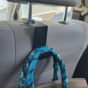 May include: A black car seat headrest hook with a blue and green patterned scarf hanging from it. The hook is attached to a gray car seat. A brown purse is visible below the scarf.
