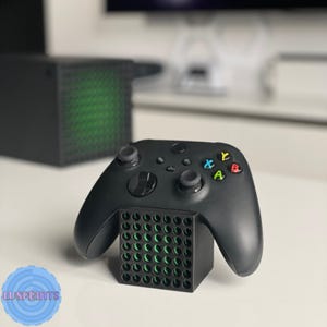 May include: Black Xbox controller resting on a black stand with green accents. The controller has colored buttons and two joysticks. A black Xbox console is visible in the background.