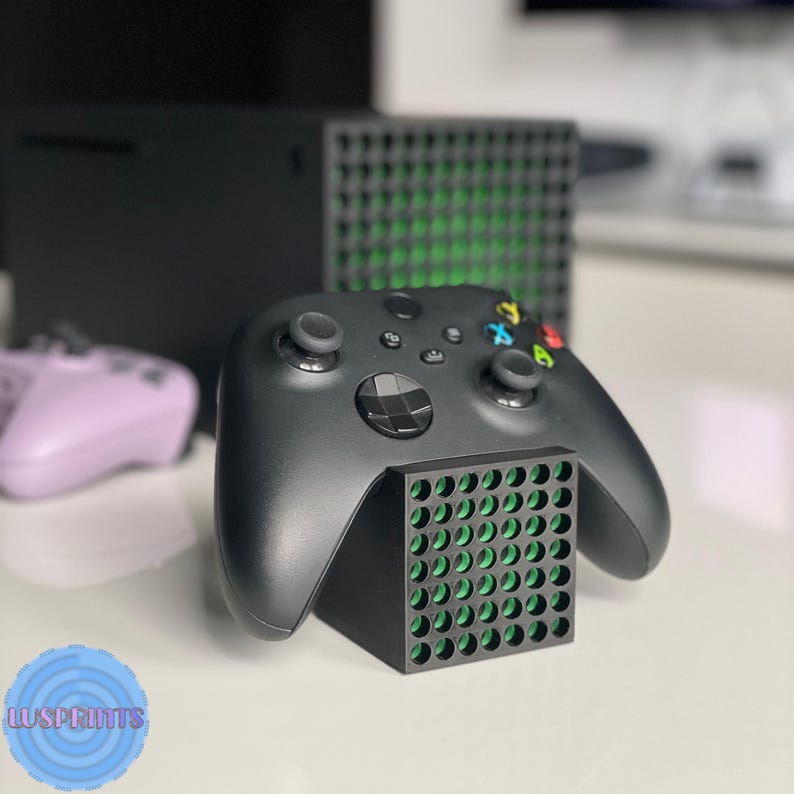 May include: Black Xbox controller resting on a black stand with green accents. A black Xbox Series X console is in the background, along with a purple controller. The stand has a grid-like design.