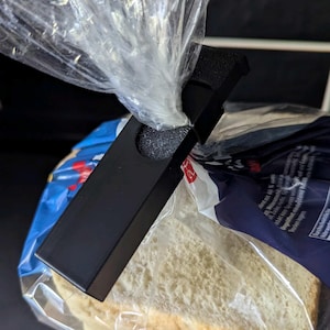 May include: A black bread clip is attached to a clear plastic bag containing sliced white bread. The clip is rectangular with a circular cutout and a textured interior. The bread is in a blue and white bag.