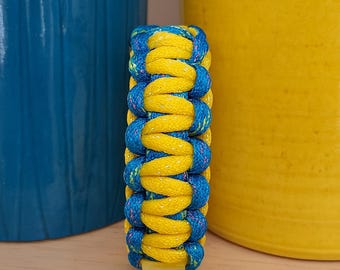 Può includere: Un braccialetto intrecciato giallo e blu, con un motivo ripetuto di corda gialla e blu. Il braccialetto è realizzato in materiale spesso e resistente. Lo sfondo include oggetti decorativi blu e gialli.