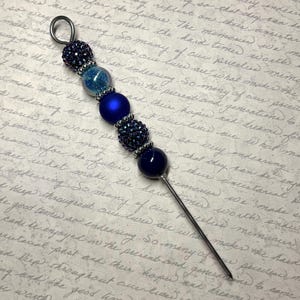 Puede incluir: Un alfiler de sombrero decorativo azul marino con un diseño de cuentas. El alfiler presenta una serie de cuentas azul marino, incluyendo elementos brillantes y texturizados, y un lazo en la parte superior. El alfiler mide aproximadamente 10 cm de largo.