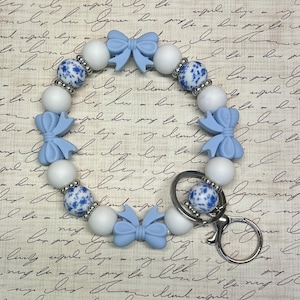 Op de afbeelding: Een sleutelhanger van witte en blauwe kralen met een zilveren sleutelring. De kralen zijn wit met blauwe bloemenpatronen en blauwe strikken.