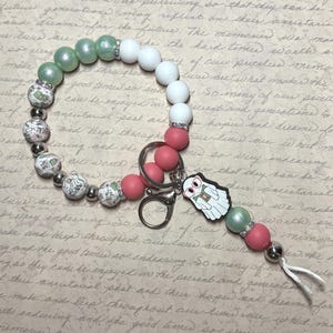 May include: A beaded keychain wristlet in shades of green, white, and pink. The wristlet features a ghost charm wearing glasses and holding a book. The beads are accented with silver-colored metal beads and rhinestones.