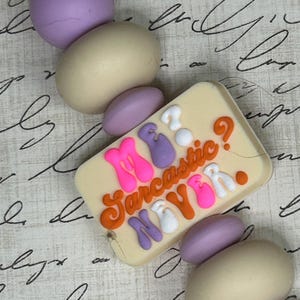 Peut inclure: Un bracelet en perles colorées avec un charme qui dit "My Sarcastic New Year." Le charme est blanc avec des lettres roses, violettes et orange. Les perles sont crème et violettes.