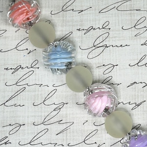 Puede incluir: Una cadena de cuentas de vidrio transparente de varios colores, incluyendo rosa, azul, morado y blanco. Las cuentas están dispuestas en línea y separadas por pequeños separadores transparentes.