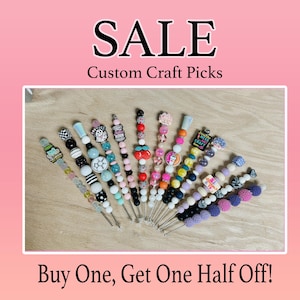 Puede incluir: Una venta de pinchos artesanales personalizados. Los pinchos están hechos con cuentas y abalorios coloridos. El texto "Buy One, Get One Half Off!" está en la parte inferior de la imagen.