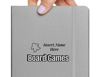 Könnte beinhalten: Ein silbernes Notizbuch mit einem schwarzen Gummibandverschluss und einem schwarz-weißen Brettspielsymbol und dem Text "Name hier einfügen Brettspiele".