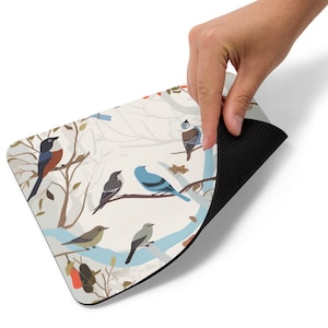 May include: A white mouse pad with a pattern of stylized birds on branches. The birds are in shades of blue, green, brown, and red. The background is a light cream color. The mouse pad has a black rubber backing.