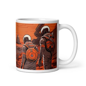 May include: White ceramic mug with a colorful illustration of two astronauts in orange spacesuits walking on a red planet. The astronauts are wearing helmets and carrying backpacks. The background is a red planet with a building in the distance.
