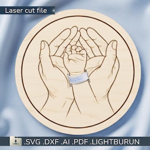 May include: A laser-cut wooden circle features a detailed outline of two adult hands cradling a baby's hand. A band around the baby's wrist reads "Baby Boy." The text "Laser cut file" is at the top. The bottom text includes file formats: .SVG .DXF .AI .PDF .LIGHTBURUN.
