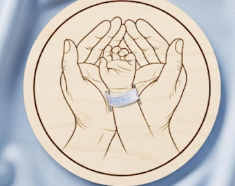 May include: A laser-cut wooden circle features a detailed outline of two adult hands cradling a baby's hand. A band around the baby's wrist reads "Baby Boy." The text "Laser cut file" is at the top. The bottom text includes file formats: .SVG .DXF .AI .PDF .LIGHTBURUN.