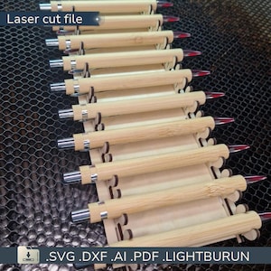 以下が含まれることがあります： 銀色のアクセントと赤い先端が付いた竹製のペンが並んでいます。ペンはレーザーカットされた木製の構造で固定されています。画像には「Laser cut file」というテキストとファイル拡張子が含まれています。