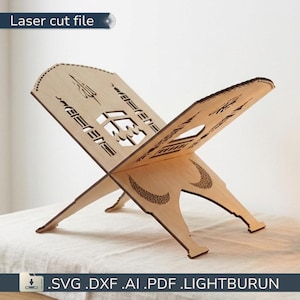 May include: A light-coloured wooden book stand with intricate laser-cut designs. The stand is in an open, angled position, with decorative patterns and the text "Laser cut file" visible. The base includes the file types: .SVG .DXF .AI .PDF .LIGHTBURUN.