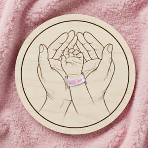 May include: A round wooden plaque with a detailed engraving of two hands cradling a baby's hand. The baby's hand has a pink band that reads "Baby Girl". The plaque is set against a soft, pink, textured background.