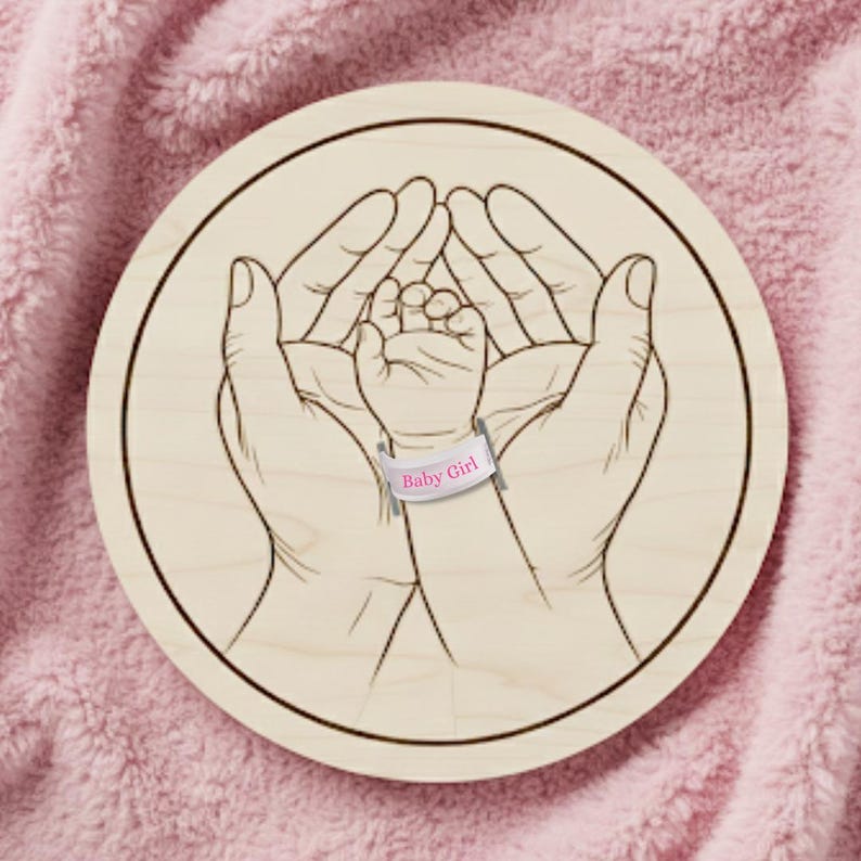 May include: A round wooden plaque with a detailed engraving of two hands cradling a baby's hand. The baby's hand has a pink band that reads "Baby Girl". The plaque is set against a soft, pink, textured background.