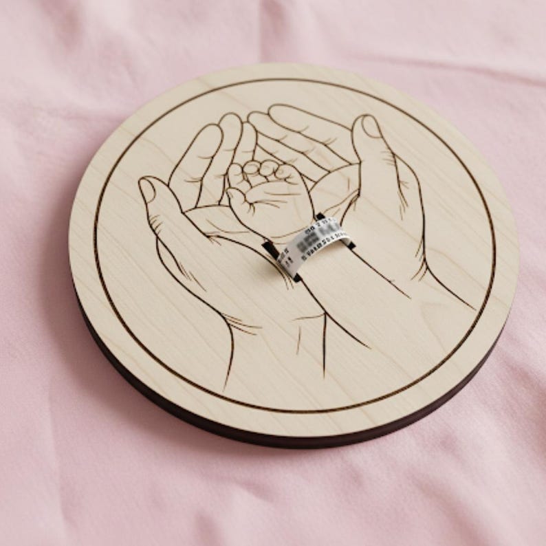 May include: A round wooden keepsake featuring an etched design of hands cradling a baby's hand. The baby's wrist has a hospital band. The wood has a light color with a dark brown border. The background is a soft pink.