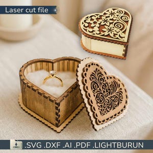 May include: A heart-shaped wooden ring box with a gold ring inside, and a separate lid with a floral design. The box is made of light brown wood and has a decorative laser-cut design. The text "Laser cut file" is visible.