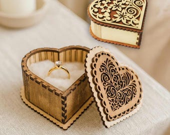 May include: A heart-shaped wooden ring box with a gold ring inside, and a separate lid with a floral design. The box is made of light brown wood and has a decorative laser-cut design. The text "Laser cut file" is visible.