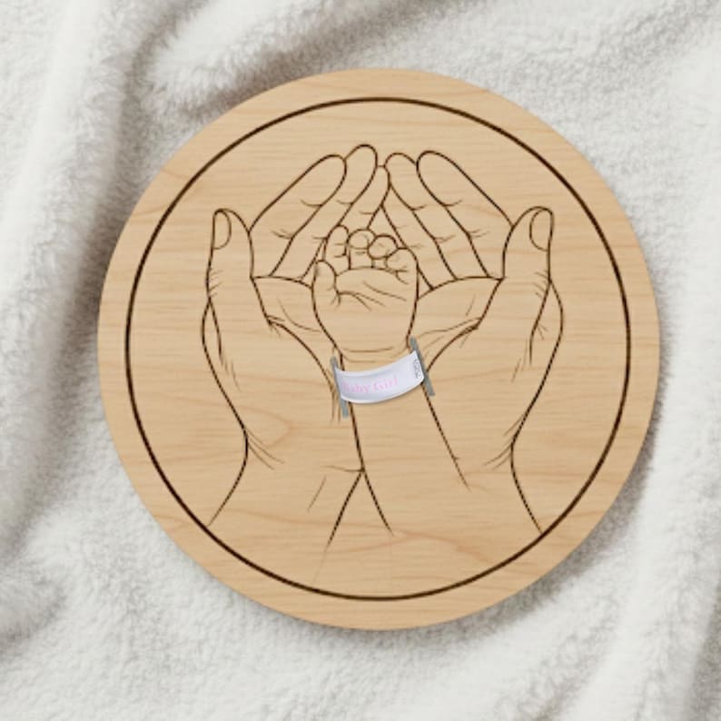 May include: A round wooden plaque with a laser-etched design. The design features two hands cradling a baby's hand, with a hospital bracelet that reads "Baby Girl". The plaque is light brown.