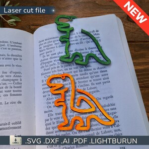 Puede incluir: Marcadores de dinosaurios cortados con láser en verde y naranja, colocados en un libro. Los marcadores son de madera y tienen forma de dinosaurio. El libro está abierto en una página con texto en francés.