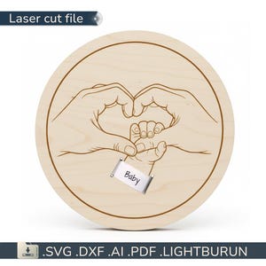 Może przedstawiać: Okrągły drewniany plik do cięcia laserowego przedstawiający kształt serca utworzony przez dłonie dorosłych trzymające dłoń dziecka. Słowo "Baby" znajduje się na małej srebrnej etykiecie. Tekst "Laser cut file" znajduje się u góry.