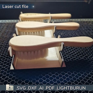 Op de afbeelding: Drie houten verzorgingsborstels met lichtgekleurde borstelharen worden getoond. De borstels hebben een gebogen handvat en staan in een houten standaard. De tekst "Laser cut file" staat linksboven. De onderkant van de afbeelding toont bestandstype-opties.