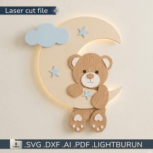 Può includere: Una lampada da parete con una mezzaluna color crema, una nuvola azzurra e tre stelle azzurre. Un orso in legno marrone con muso bianco e cuscinetti a forma di cuore è posizionato davanti alla luna. Il testo recita Laser cut file.