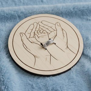 May include: A round wooden plaque with a laser-etched design of adult hands cradling a baby's hand. A hospital wristband is on the baby's wrist. The plaque is light brown with a dark brown edge, set against a blue background.