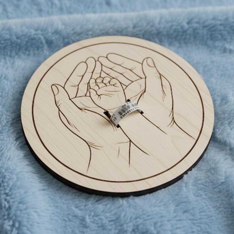 May include: A round wooden plaque with a laser-etched design of adult hands cradling a baby's hand. A hospital wristband is on the baby's wrist. The plaque is light brown with a dark brown edge, set against a blue background.