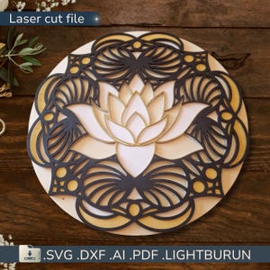 Peut inclure: Fichier de découpe laser d'un motif de fleur de lotus en couches. La fleur est blanche et dorée, entourée d'un motif de filigrane noir et doré. Le motif est sur une base en bois ronde. Le texte en haut indique "Laser cut file".