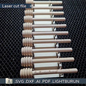 May include: A collection of wooden honey dippers, each with a grooved head and a long handle. The dippers are arranged in a vertical row, resting on a light-colored wooden base. The text "Laser cut file" is visible at the top.