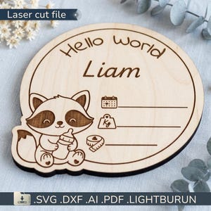 Op de afbeelding: Houten baby aankondigingsbord met de tekst "Hello world Liam". Het bord toont een cartoon wasbeer met een fles, met ruimte voor datum, gewicht en lengte. De woorden "Laser cut file" staan linksboven.