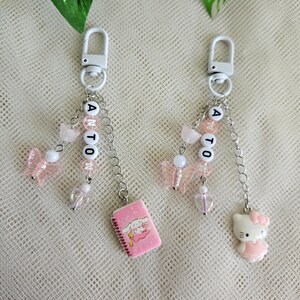 May include: Two pink and white charm keychains with a Hello Kitty charm, a pink notebook charm, and pink butterfly charms. The keychains spell out the name "ANTON" in white letters.