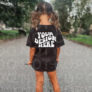 Könnte beinhalten: Ein schwarzes T-Shirt mit dem Text "YOUR DESIGN HERE" in weißer Schrift im Retro-Stil. Das Shirt wird von einem Kind getragen, das auf einer gepflasterten Straße steht.