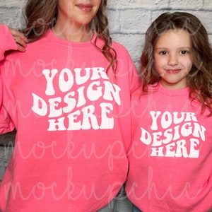 Könnte beinhalten: Zwei rosa Sweatshirts mit dem Text "YOUR DESIGN HERE" in weißer, retro-artiger Schrift. Die Sweatshirts werden von zwei Personen getragen, einem Erwachsenen und einem Kind.