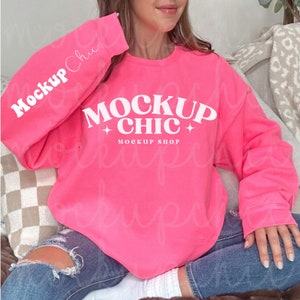 以下が含まれることがあります： 明るいピンクのトレーナーに、白い文字で「MOCKUP CHIC」と書いてあり、「CHIC」の両側に星が1つずつ付いています。「MOCKUP SHOP」という文字がメインの文字の下にあります。トレーナーは、ソファに座っている人が着ています。