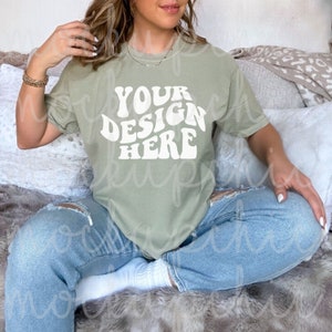 May include: A woman wearing a light green t-shirt with the text "YOUR DESIGN HERE" in white. She is sitting on a fuzzy rug with her legs crossed.