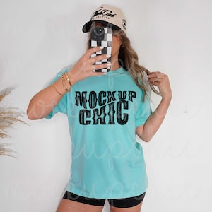 May include: A light blue t-shirt with the text "MOCKUP CHIC" in black. The person in the image is wearing a tan baseball cap and black shorts. The person is holding a phone with a black and white checkered case.