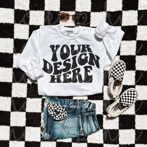 May include: White sweatshirt with the text "YOUR DESIGN HERE" in black retro font, laying on a black and white checkered surface with a pair of black and white checkered slip-on shoes, blue denim shorts, and sunglasses.