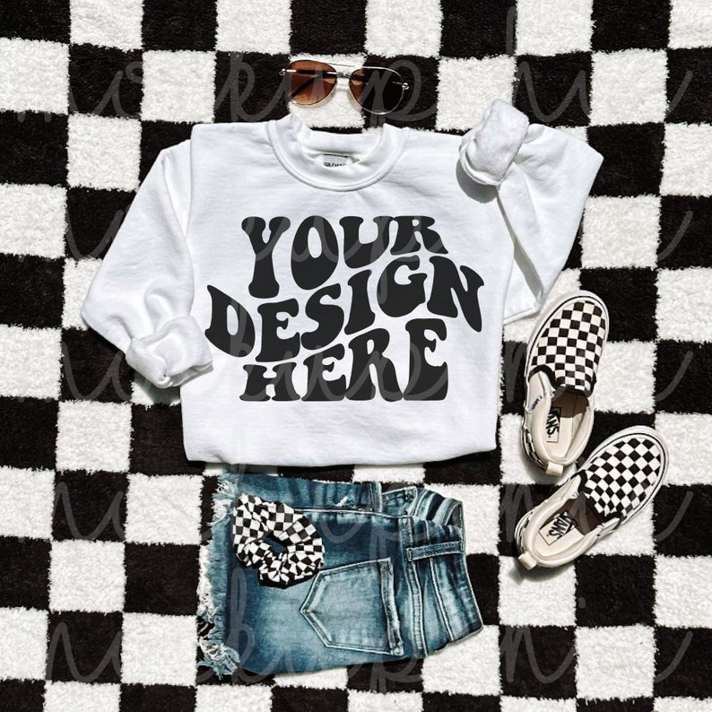 May include: White sweatshirt with the text "YOUR DESIGN HERE" in black retro font, laying on a black and white checkered surface with a pair of black and white checkered slip-on shoes, blue denim shorts, and sunglasses.