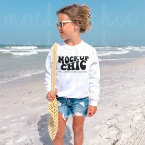 Może przedstawiać: Biała bluza z napisem "MOCKUP CHIC" w kolorze czarnym, w połączeniu z dżinsowymi szortami. Bluza ma długie rękawy i okrągły dekolt. Żółta szydełkowa torba jest przewieszona przez ramię. Tłem jest scena na plaży.