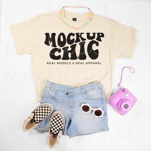 Könnte beinhalten: Cremefarbenes T-Shirt mit dem schwarzen Text "MOCKUP CHIC", kombiniert mit hellblauen Jeansshorts, karierten Slip-On-Sneakers und einer rosa Kamera. Eine Blumenkette und weiße Sonnenbrillen vervollständigen den Look.