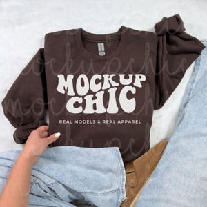 May include: A brown sweatshirt with the text "MOCK UP CHIC" in white, stylized lettering. The text is in a retro font style. The sweatshirt is worn by a person wearing blue jeans and brown boots. The text "REAL MODELS & REAL APPAREL" is below the main text.