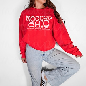 Op de afbeelding: Een rode sweatshirt met de witte tekst "MOCKUP CHIC" en de tekst "NO AI Real Models & Real Apparel". De sweatshirt wordt gecombineerd met lichtblauwe jeans en witte sneakers.
